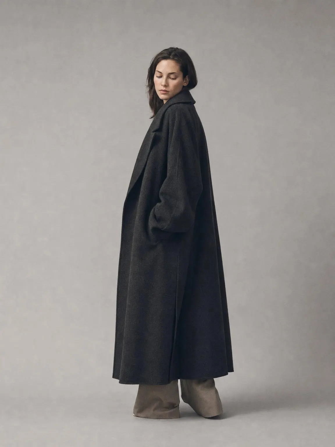 A model wearing an oversized charcoal wool coat against a pale grey studio backdrop.